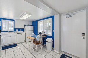 Een compacte keuken met witte kasten, een dubbele spoelbak, een koelkast en een eethoek met een stoel en tafel.