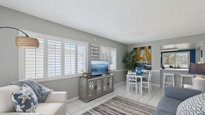 A living room with a television, seating, and a dining area with chairs and a table. - Chateau by the Sea 10B- Oceanfront Paradise (New Smyrna Beach)