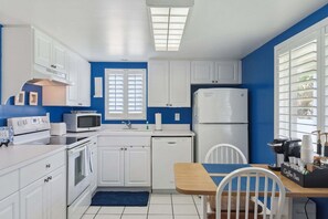 A compact kitchen with white cabinets, a microwave, a stove, a refrigerator, and a coffee bar setup. - Chateau by the Sea 10B- Oceanfront Paradise (New Smyrna Beach)