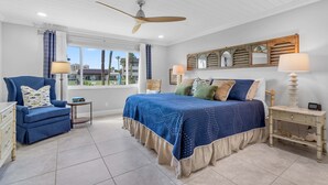 A bedroom with a large bed, a ceiling fan, a window with curtains, a dresser, and a nightstand with a lamp. - Chateau by the Sea 12D Ocean Breeze & Views! (New Smyrna Beach)