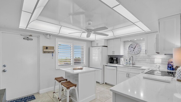 A modern kitchen with white cabinetry, a central island, and a ceiling fan. - Chateau by the Sea 12D Ocean Breeze & Views! (New Smyrna Beach)