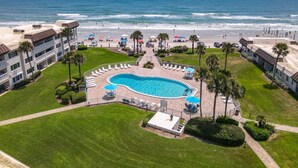 A resort with a swimming pool surrounded by palm trees, a beach, and ocean views. - Chateau by the Sea 34D Life's Better by the Sea! (New Smyrna Beach)