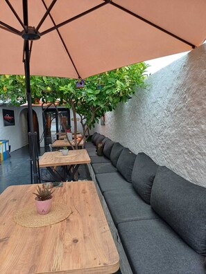 Un área de estar al aire libre con mesas de madera y un banco con cojines grises, rodeada de follaje y una pared con textura.