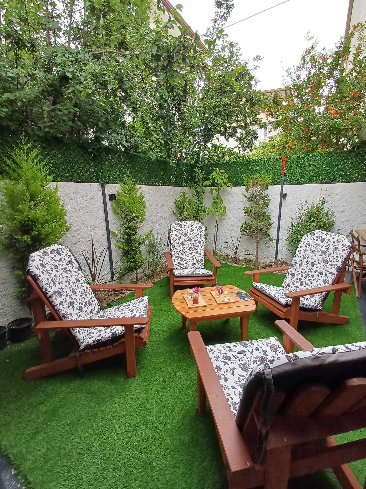Un patio con mobiliario de madera y césped artificial, rodeado de un muro blanco y follaje.