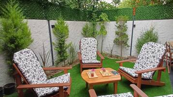 Un patio con mobiliario de madera y césped artificial, rodeado de un muro blanco y follaje.