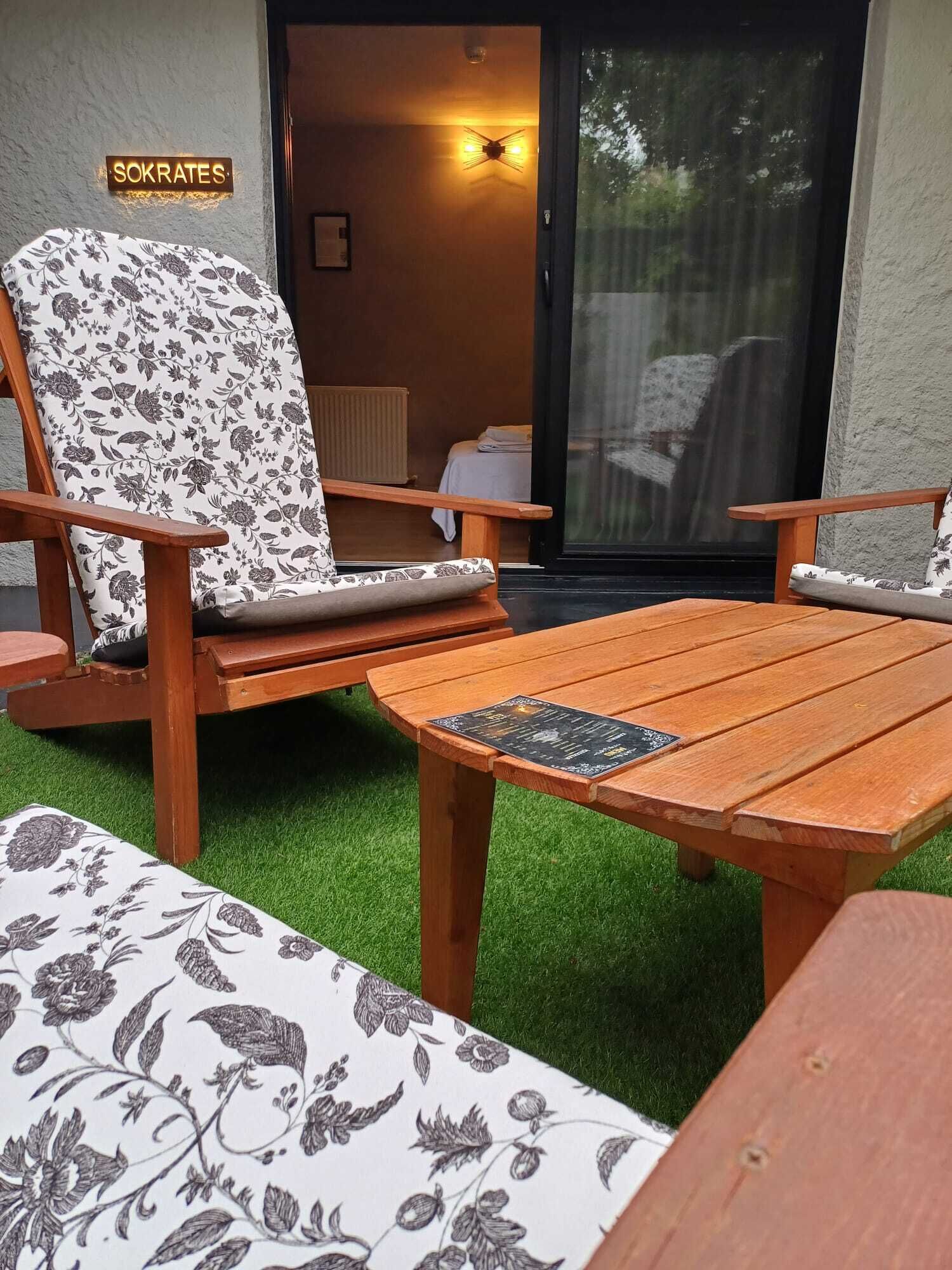 A patio area with a wooden table and two chairs with floral upholstery. A sign reading "SOKRATES" is visible on the wall.