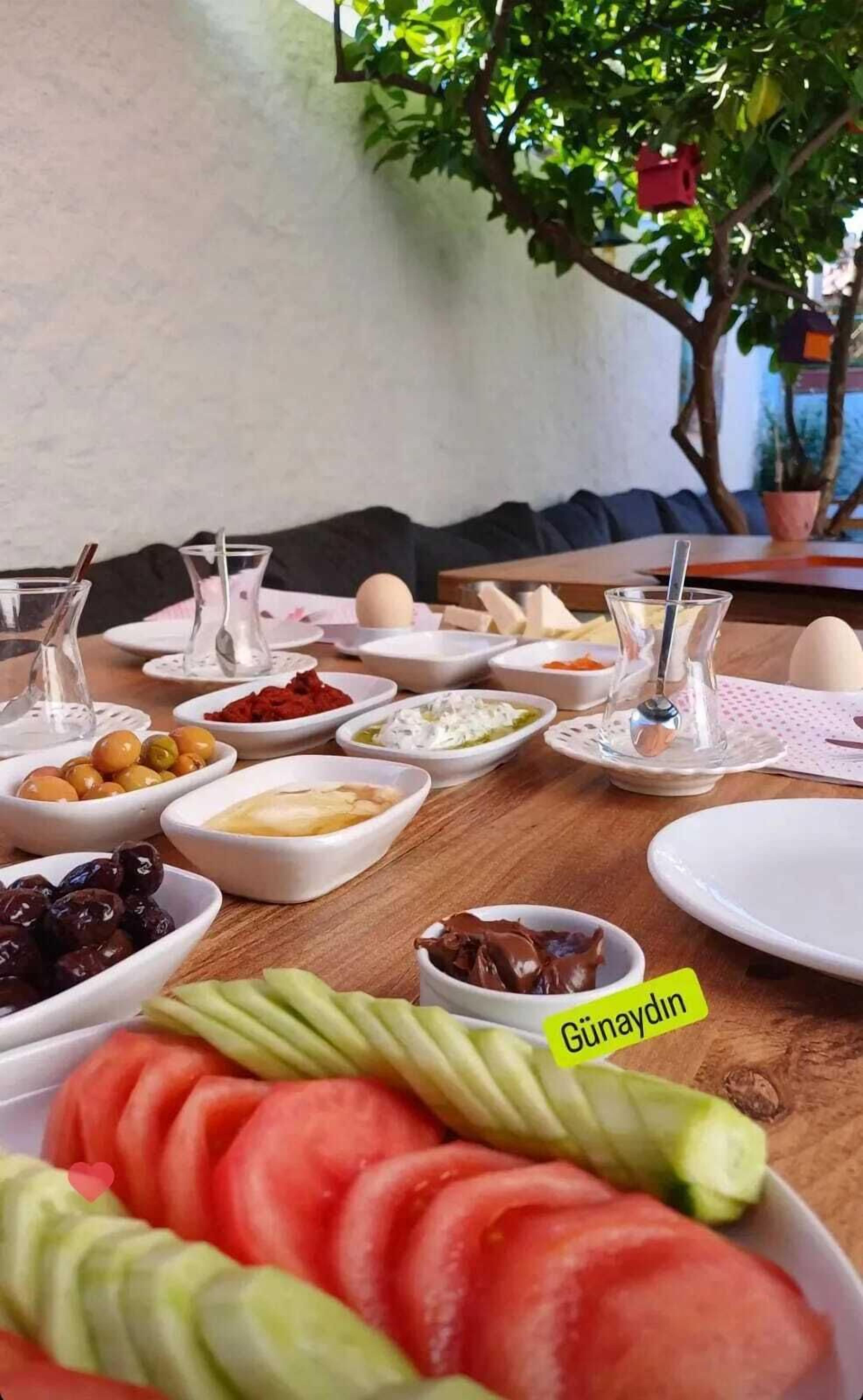 Une table dressée pour un repas, avec différents plats, notamment un saladier de tomates et de concombres coupés en tranches, et une assiette portant l’inscription « Günaydın ».