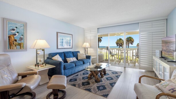 A living room with a blue sofa, a glass coffee table, and a view of the ocean through large sliding doors. - Beachfront Bliss at Chateau by the Sea 24C (New Smyrna Beach)