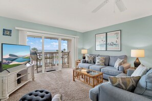 A living room with a TV, sofa, coffee table, and a view of the outdoors. - Chateau by the Sea 35C Where Coastal Meets Cozy (New Smyrna Beach)