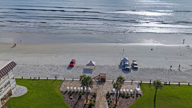 A beach view with a building, a parking area, and a few people walking. - Chateau by the Sea 13B Ocean Views & Cozy Comfort! (New Smyrna Beach)