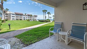 A balcony with two chairs and a small table, overlooking a well-maintained lawn and a building with multiple balconies. - Chateau by the Sea 13B Ocean Views & Cozy Comfort! (New Smyrna Beach)