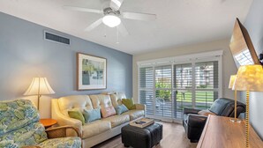 A living room with a sofa, armchair, ottoman, and a desk with a lamp. - Chateau by the Sea 13B Ocean Views & Cozy Comfort! (New Smyrna Beach)