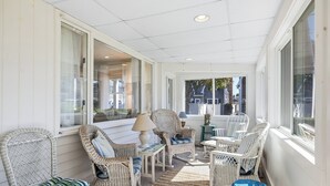 A covered porch with wicker furniture, including chairs and a table, and large windows providing views of the outside. - Recently updated home sleeping 10, steps to the beach! (Harwich Port)