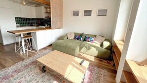 Kompakti olohuone, jossa on pieni keittiönurkkaus, vihreä sohva, puinen sohvapöytä ja kuviollinen matto.