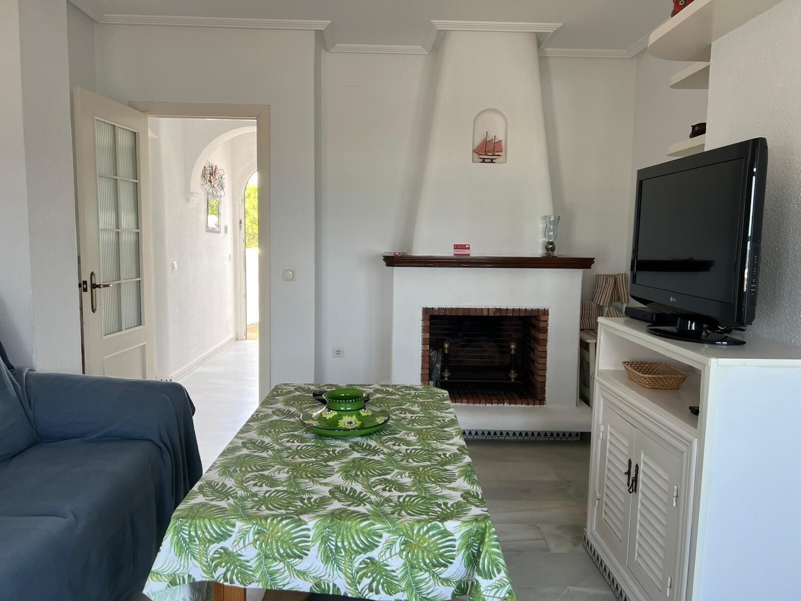 A room with a fireplace, a television, a table with a leaf-patterned tablecloth, and a blue sofa.