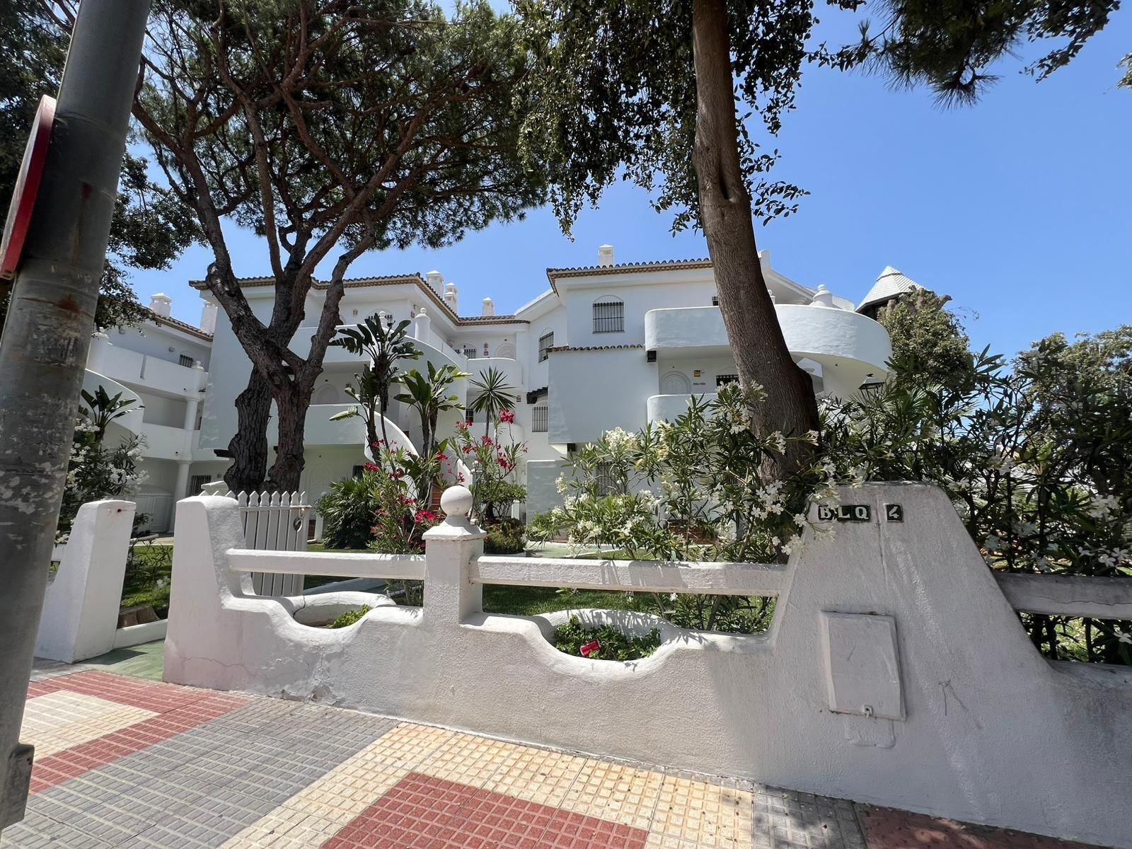 Une entrée fermée par un portail et bordée d’une clôture blanche, un chemin dallé et, au fond, un bâtiment aux murs blancs et doté de nombreux balcons.