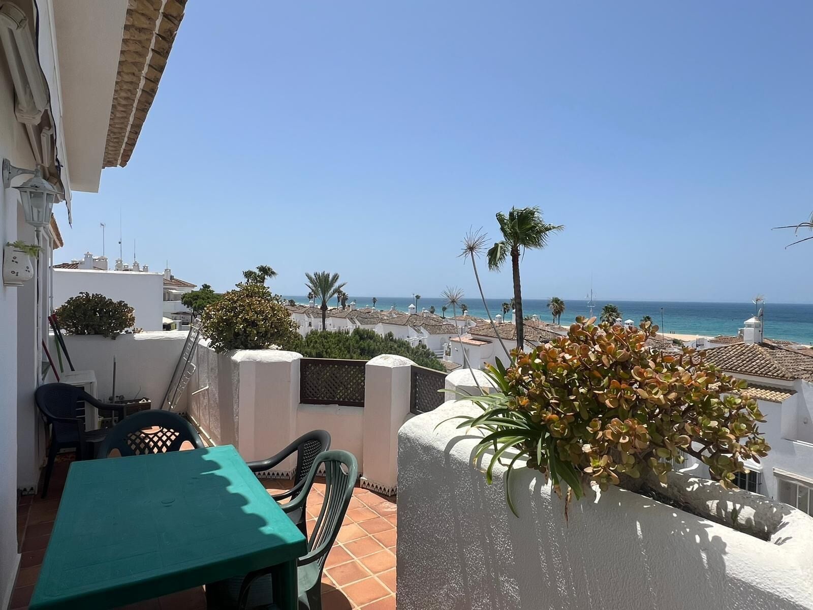 Une terrasse avec une table et des chaises vertes, un mur blanc orné d’une plante, et une vue sur la mer et les bâtiments en arrière-plan.