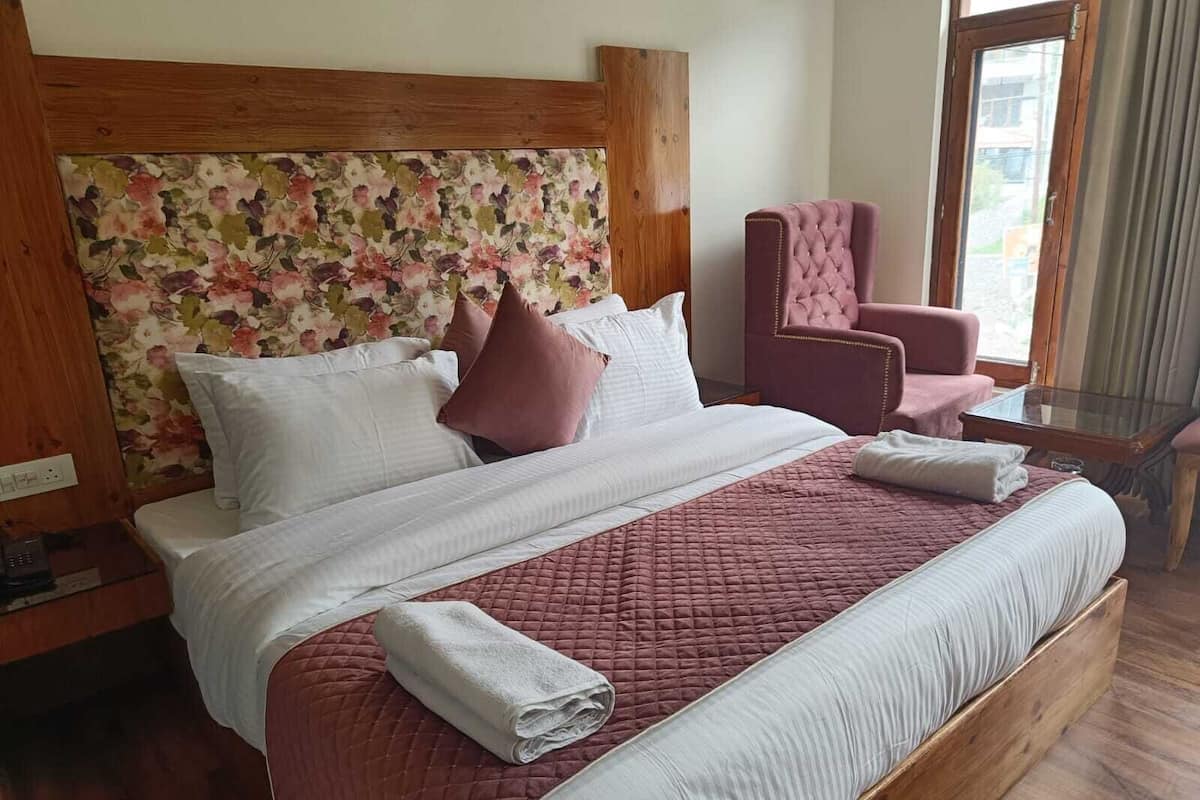 A hotel room with a large bed, a floral headboard, a pink armchair, and a glass table.