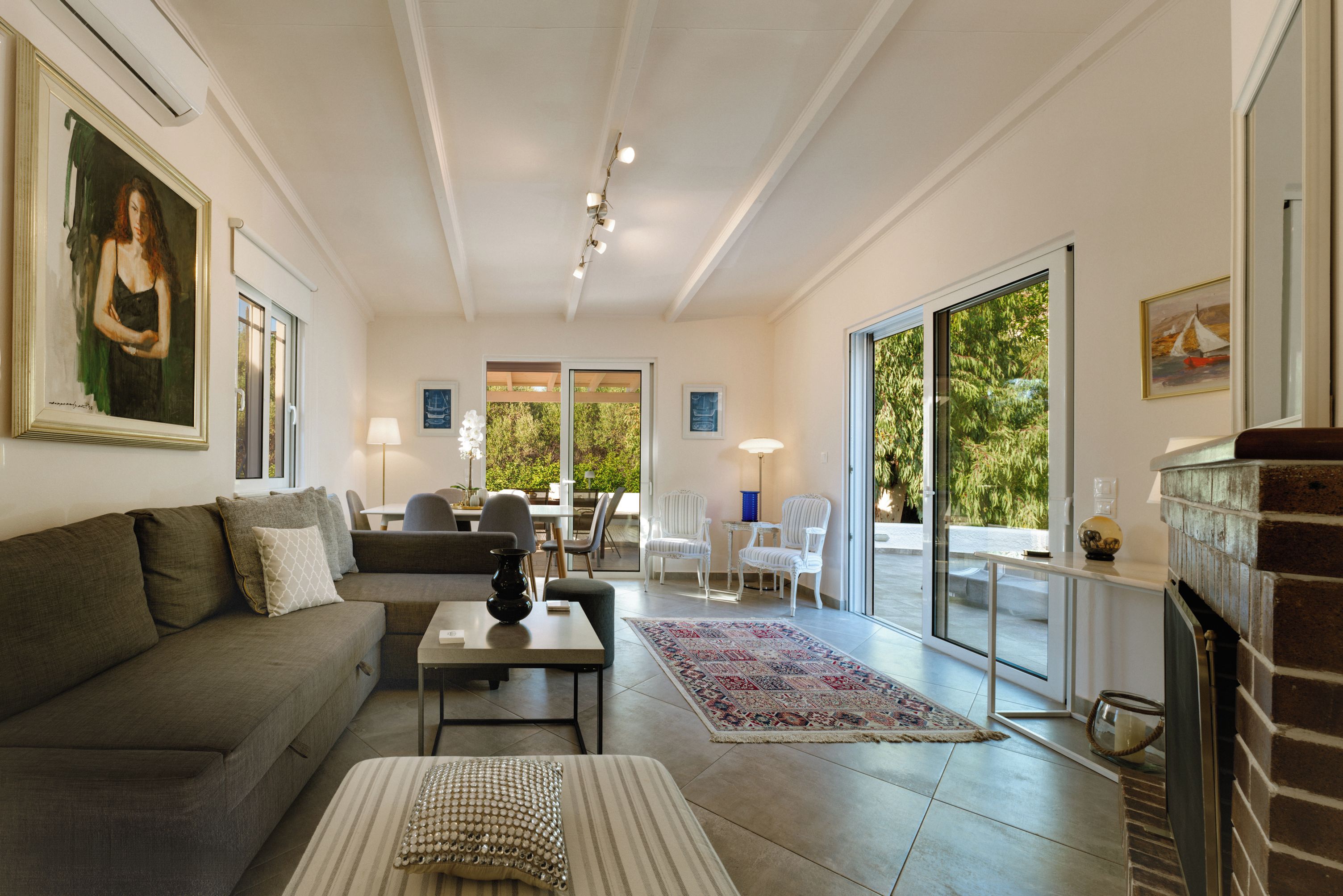 A spacious living room with a sofa, a coffee table, and a fireplace. There are two doors leading to a balcony with outdoor furniture.