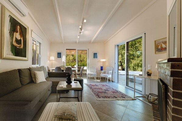 A spacious living room with a sofa, a coffee table, and a fireplace. There are two doors leading to a balcony with outdoor furniture.