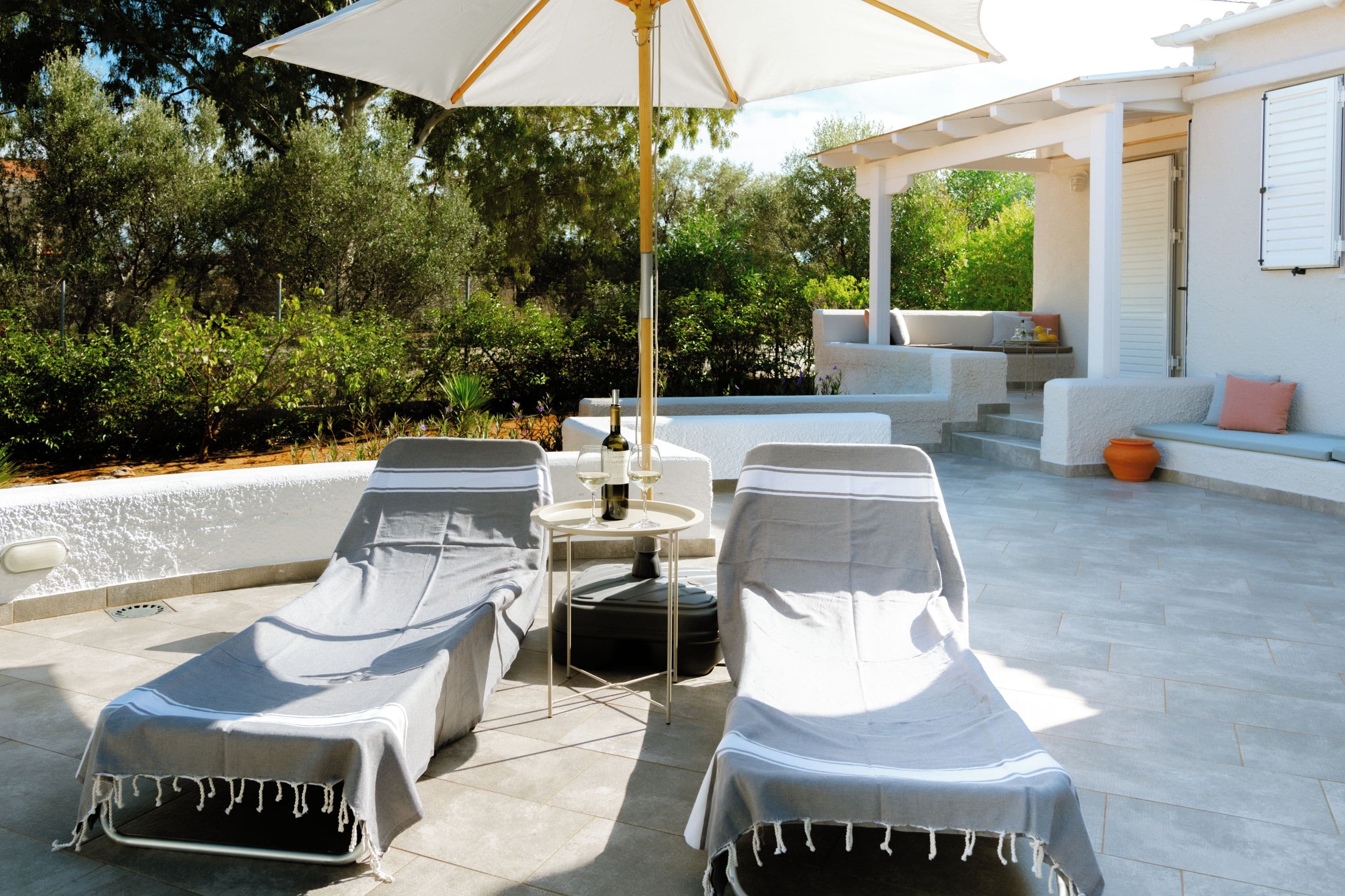 Een zonnig terras met lounge fauteuils en een tafel, omgeven door een wit gebouw en weelderige beplanting.