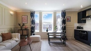 A modern living room with a sofa, coffee table, and dining area. There are large windows with curtains, a city view, and a kitchen area with cabinets and appliances. - The Balham Hideout - Trendy 2bdr Flat (London)
