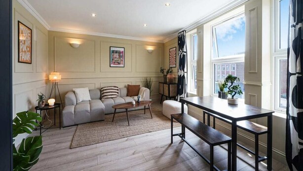 A living room with a sofa, coffee table, and a dining table. There are wall-mounted lights, a large window with curtains, and a view of the outside. - The Balham Hideout - Trendy 2bdr Flat (London)