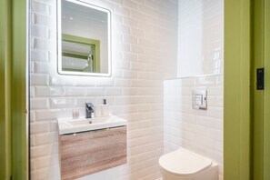 A bathroom with white tiled walls, a white sink, a mirror, and a toilet. - The Balham Place - Stylish 2bdr Flat (London)