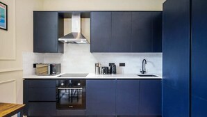 A compact kitchen with dark cabinetry, a stainless steel range hood, a microwave, a coffee maker, and a sink. - The Balham Place - Stylish 2bdr Flat (London)