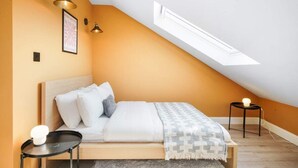 Une chambre avec un plafond en pente, un lit avec une literie blanche, un plaid à motifs, deux tables de chevet avec des lampes et un tableau encadré au mur.