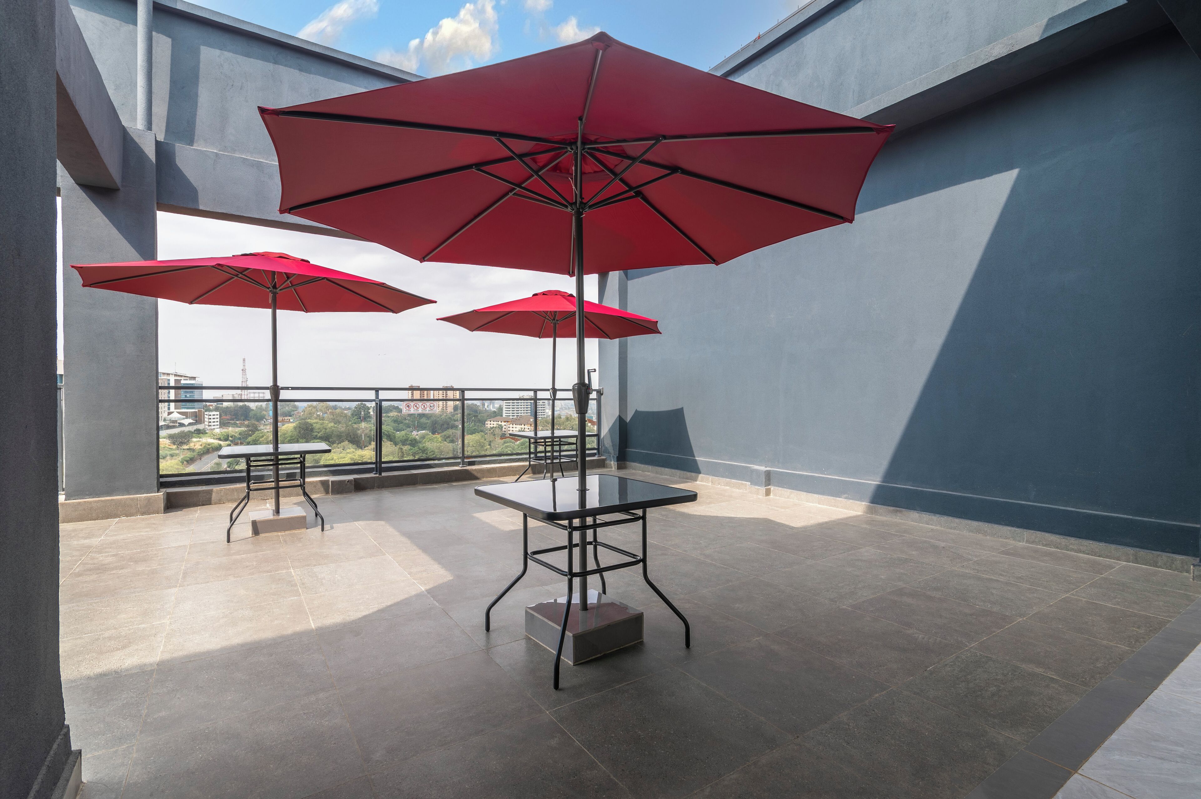 A rooftop terrace with two tables and two red umbrellas, offering a view of the cityscape.