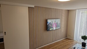 A modern living room with a flat-screen TV mounted on a wall with vertical wooden slats, a glass coffee table, and a potted plant. - Luxury Apartment Garden (Brienz)