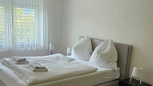A neatly made bed with white linens and pillows, a bedside table with a lamp, and a window with sheer curtains. - Luxury Apartment Garden (Brienz)