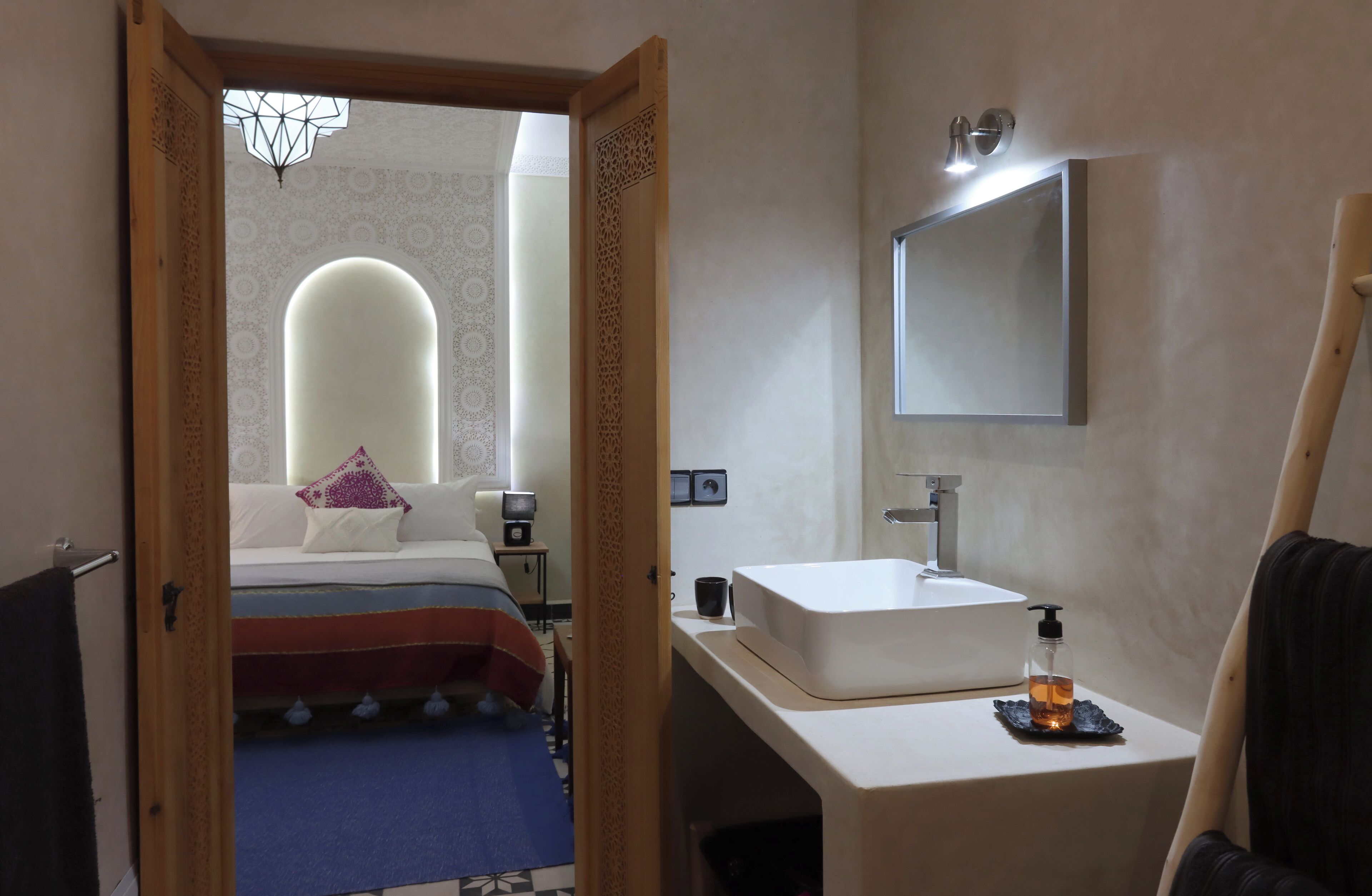 A hotel room with a bed, a bedside table, a mirror, a sink, and a towel rack.