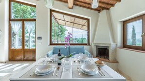 Un coin repas avec une table dressée pour quatre personnes, une cheminée et de grandes fenêtres donnant sur un paysage verdoyant.