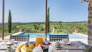 Une table à manger dressée avec des aliments pour le petit-déjeuner, donnant sur une piscine et la campagne environnante.