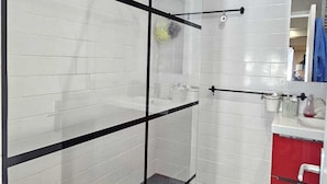 A modern bathroom with white tiled walls, a glass shower enclosure, a red cabinet, and a mirror.