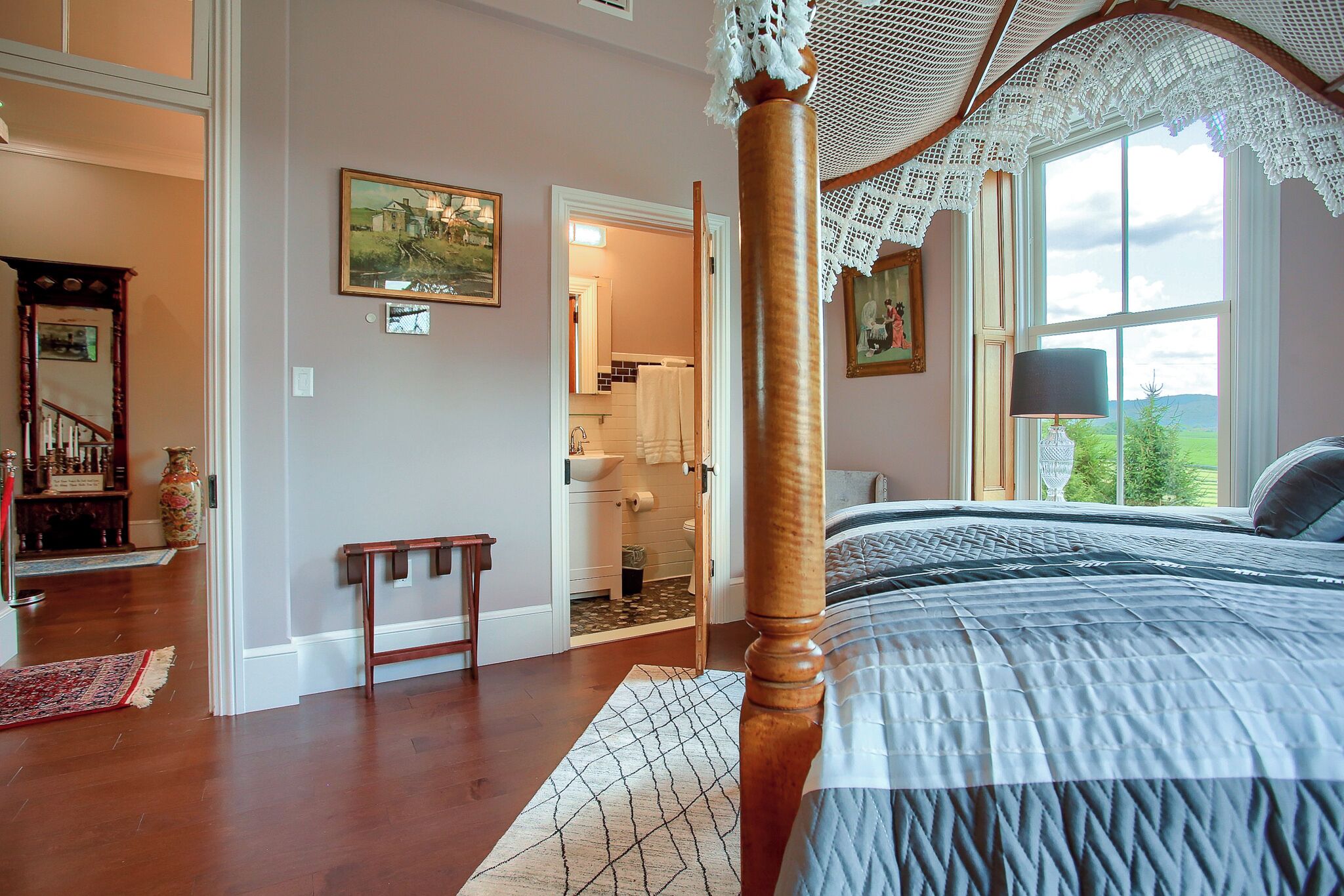 A bedroom with a four-poster bed, a view of the outdoors through a large window, and a hallway leading to a bathroom.