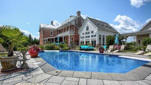 Una piscina grande con un área de patio techada, rodeada por una casa de ladrillo y una casa blanca con techo a dos aguas.