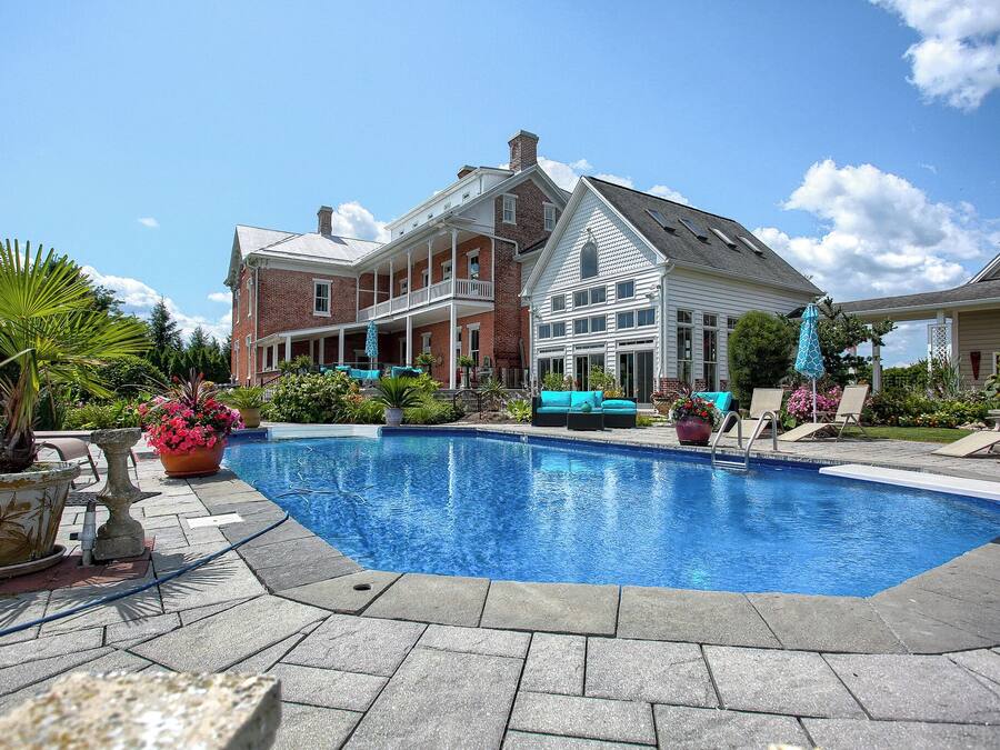 A large swimming pool with a covered patio area, surrounded by a brick house and a white house with a gabled roof.