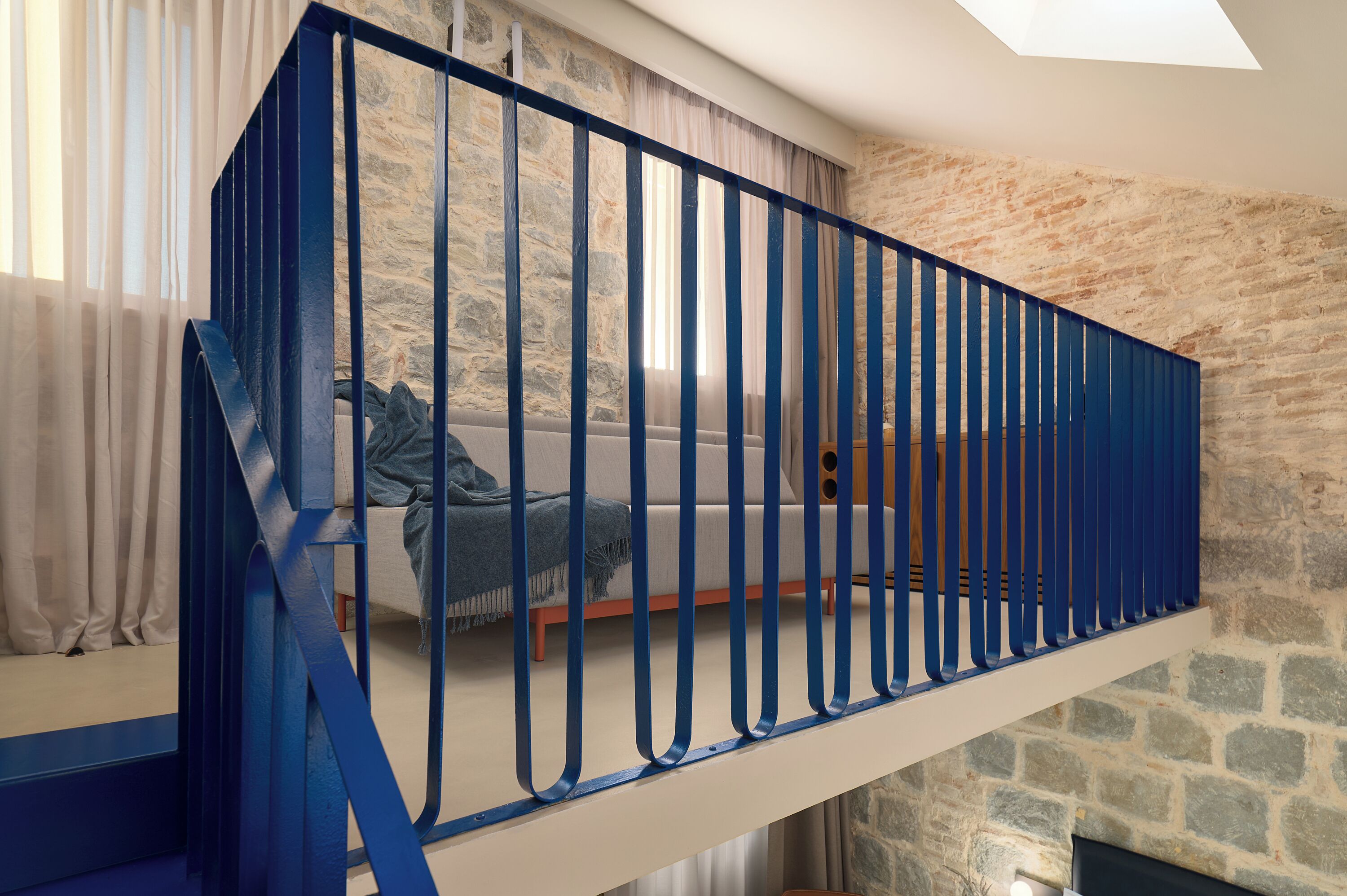 A modern loft bedroom with a stone wall, a blue railing, and a large window with sheer curtains.