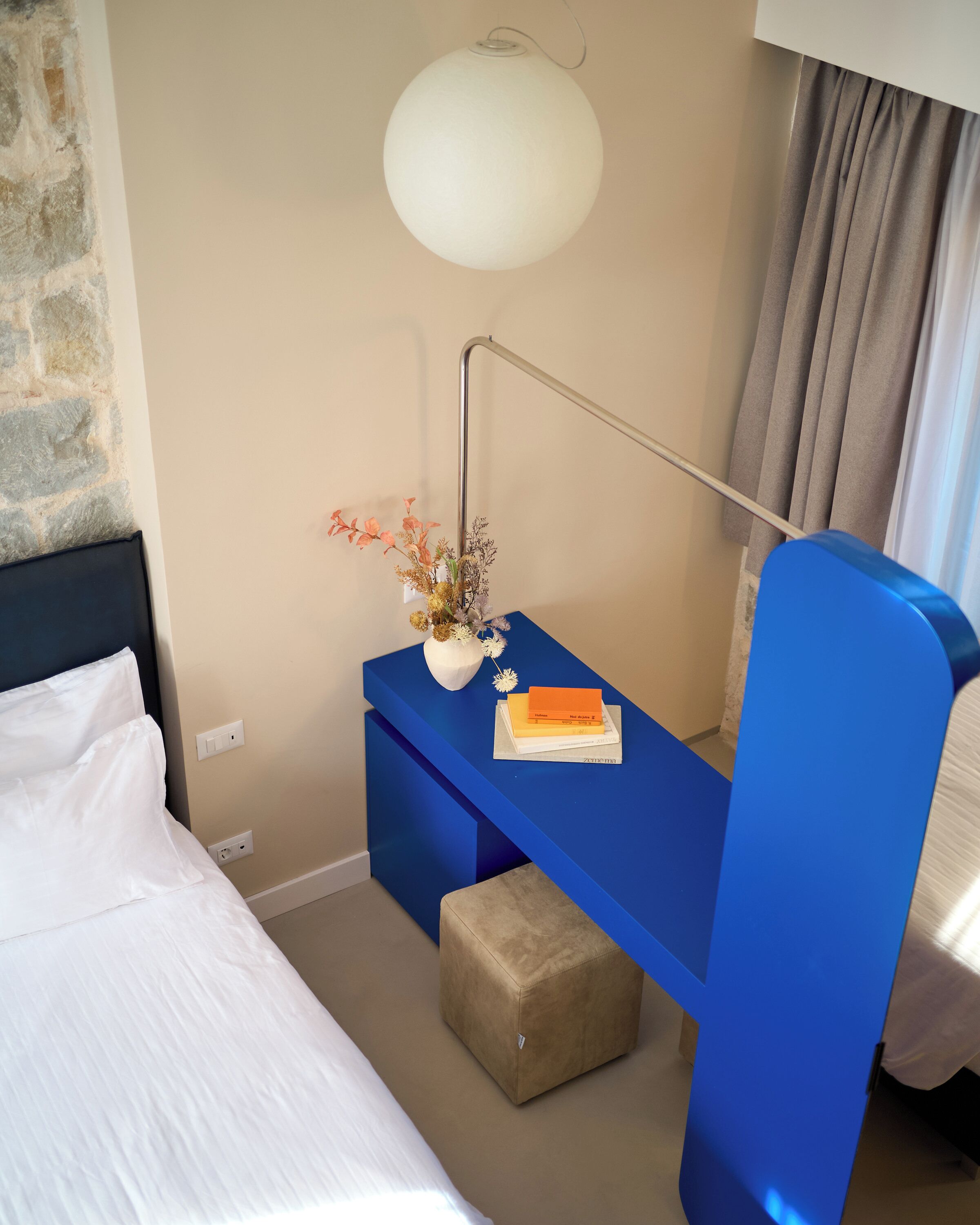 Ein Hotelzimmer mit einem Bett, einem blauen Schreibtisch mit Lampe, einer Vase mit Blumen und einem Fenster mit Vorhängen.