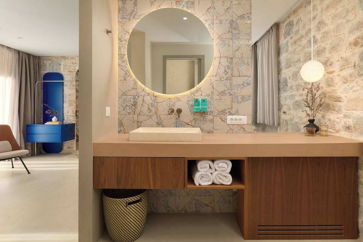 Un baño moderno con salpicadero de mármol, un espejo redondo, tocador de madera y estante para toallas.