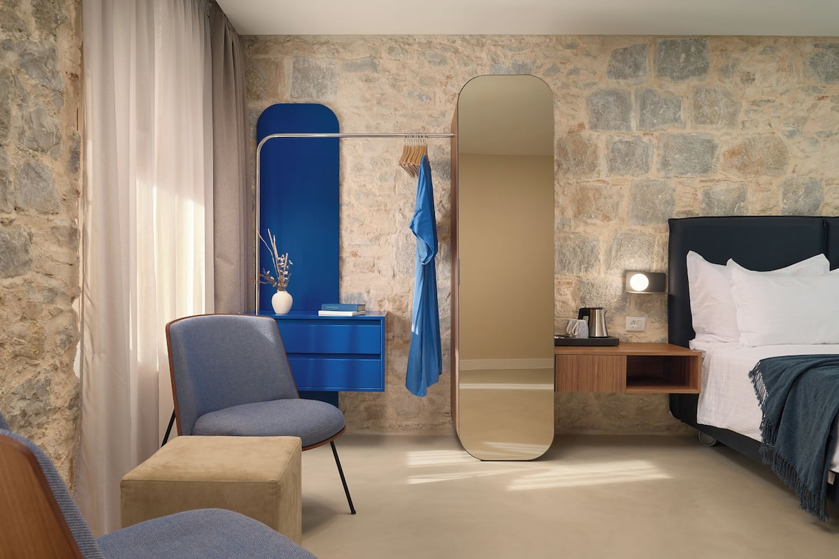 Habitación de hotel con pared de piedra, un espejo grande, una cama con ropa blanca, una mesita de noche con lámpara y una silla azul.