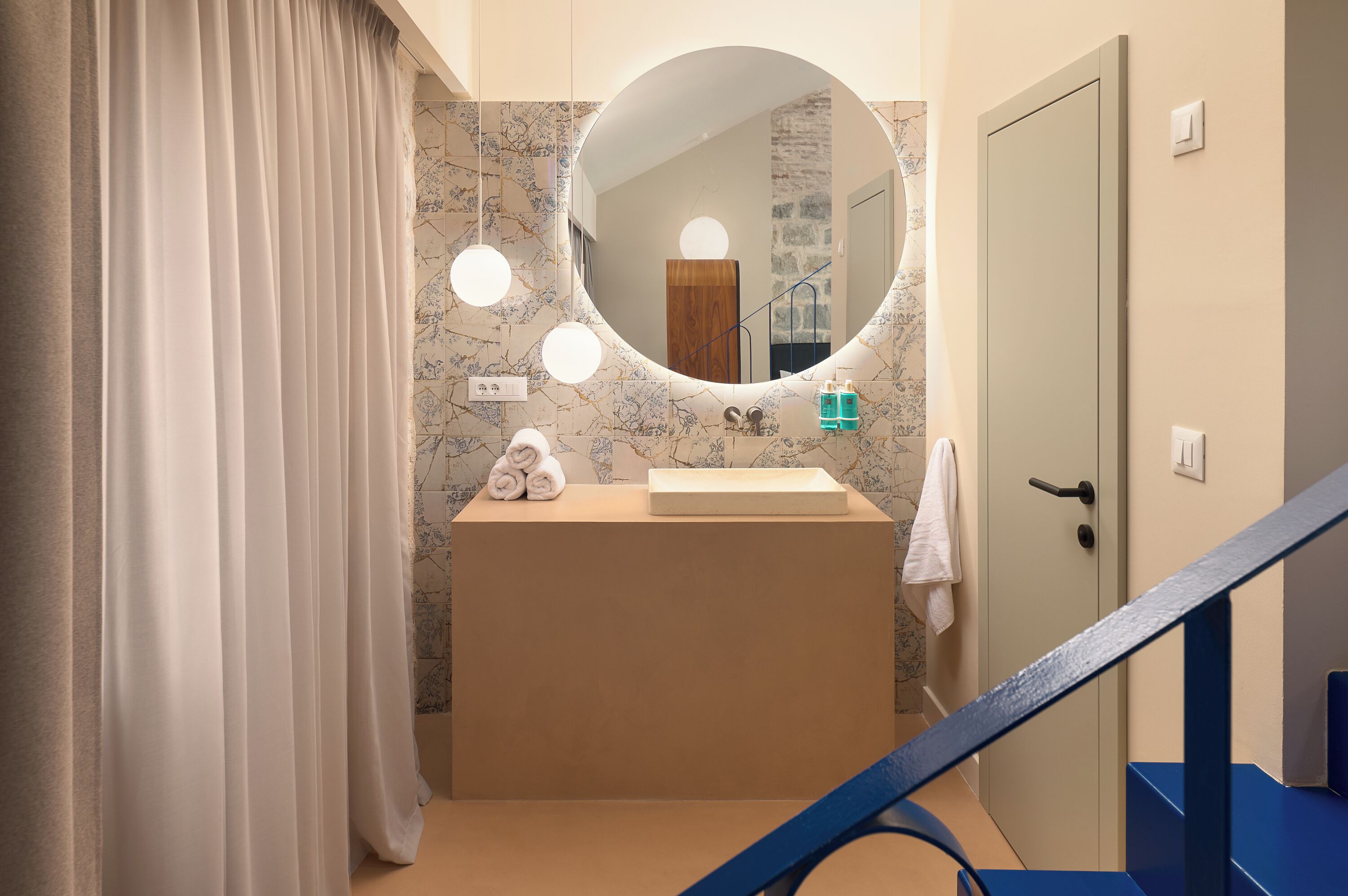 A bathroom with a marble-tiled wall, a round mirror with lights, a sink, and a towel rack.