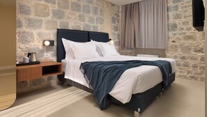 Un dormitorio con pared de piedra, una cama con ropa de cama blanca, cabecero oscuro, mesita de noche con lámpara y televisor colgado.