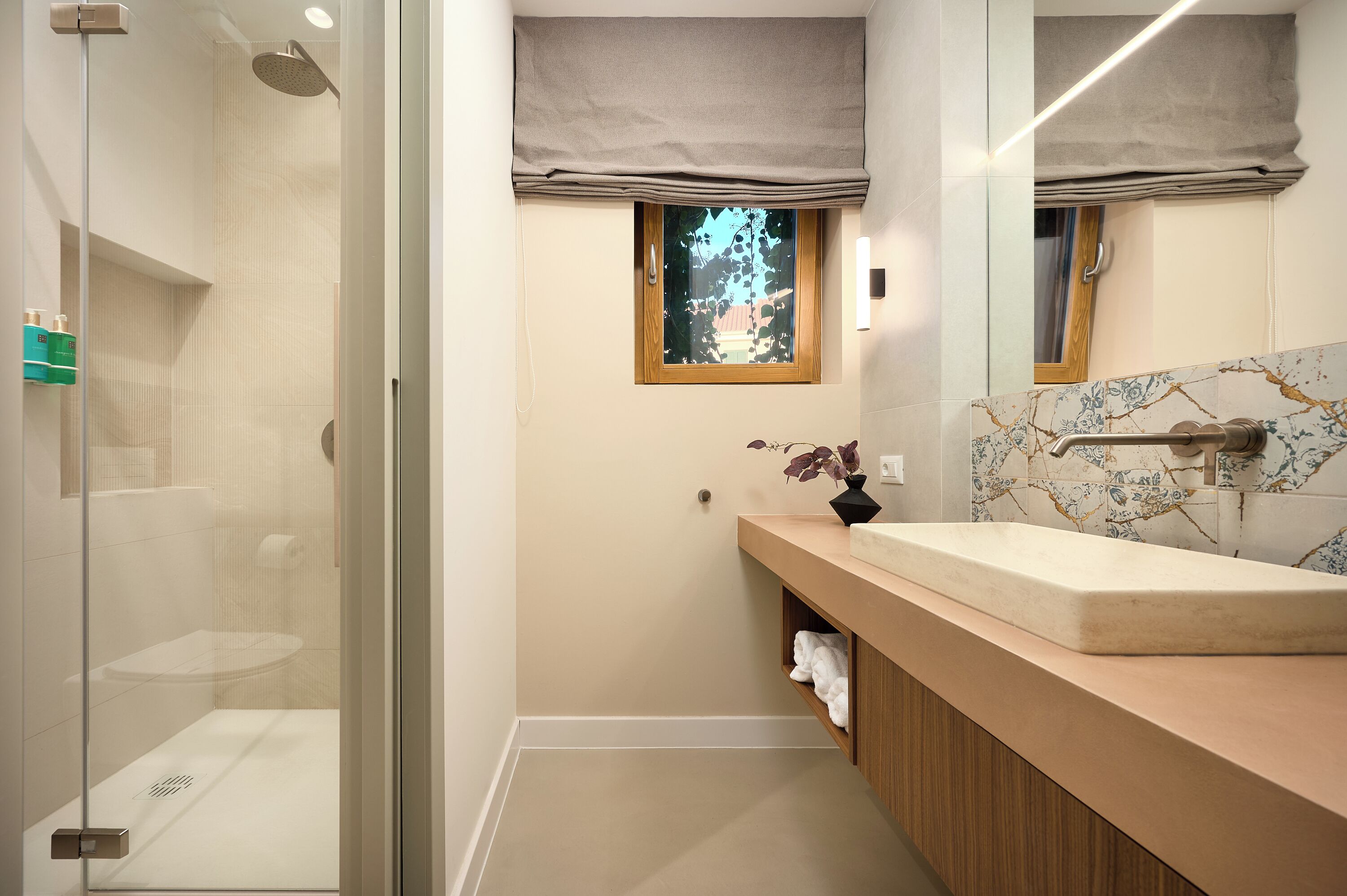 A modern bathroom with a glass shower enclosure, a wooden vanity with a marble backsplash, and a window with a view of greenery.