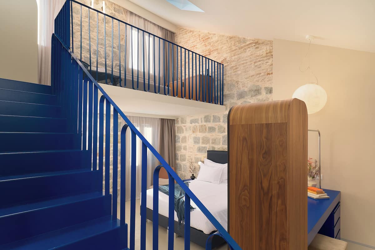 Un dormitorio con una cama doble, una mesita de noche, un escritorio de madera, una escalera azul y una pared de piedra.