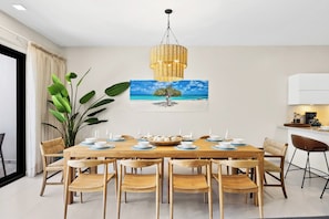 Ein Essbereich mit einem Holztisch, gedeckt für sechs Personen, umgeben von Stühlen, einer großen Pflanze und einem Bild mit einer Strandansicht.