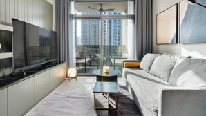 A modern hotel room with a large flat-screen TV, a grey sofa, a small round coffee table, and a balcony with a city view.